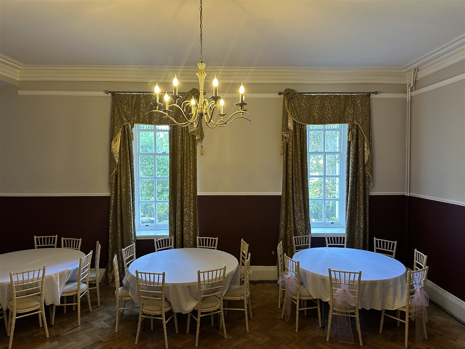 Round tables with white tablecloths and limewash chairs beneath two tall draped windows