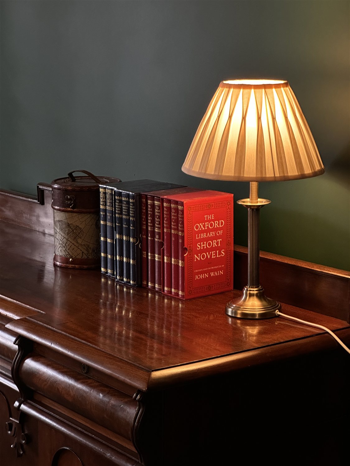 A glowing table lamp and leather-bound books on a mahogany sideboard