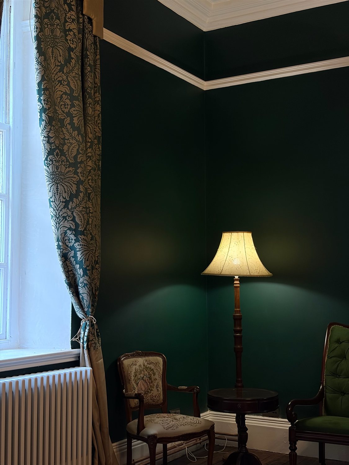 A lit floor lamp in the corner of the Green Room beside a damask curtain